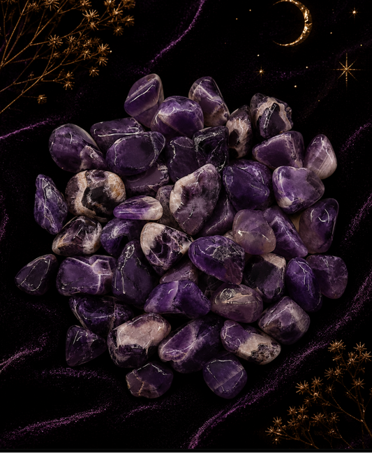A pile of polished purple amethyst tumbled stones on a dark purple velvet fabric background, decorated with gold-toned dried botanicals and a crescent moon.